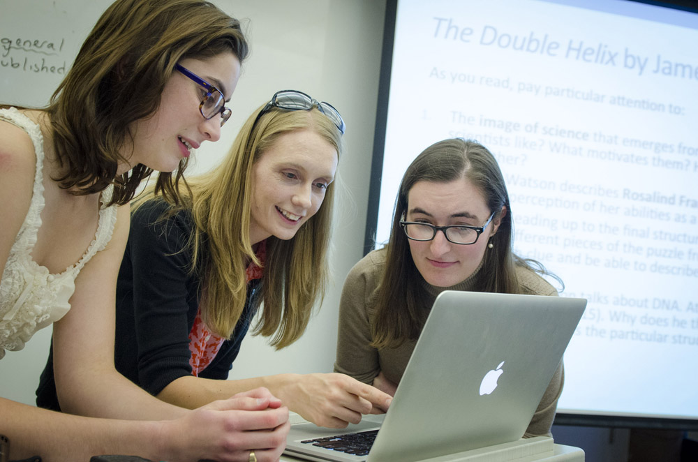 Two students with professor look at laptop.