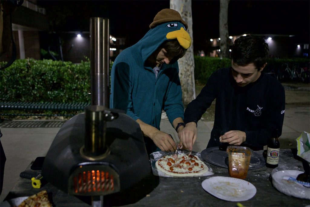 Two students place toppings on a pizza next to a pizza oven.