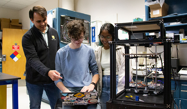 Steven Santana and two students work together on a laptop near a printing machine.