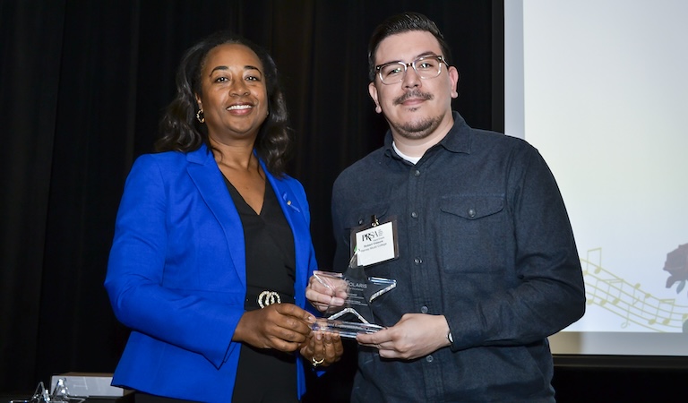 Two staff members of the Harvey Mudd communications and marketing team hold a glass award shaped like a star. They are both smiling and standing in front of a black curtain backdrop.