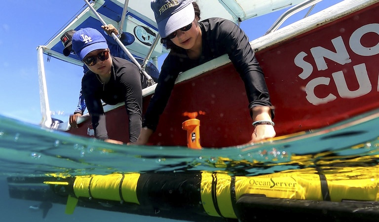 Two girls in baseball caps and sunglasses lean over the side of a red boat. They are both working on a long yellow cylinder floating in greenblue water.