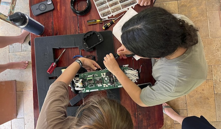 Two students work on a computer board. Screw drivers, cables and a tool box lay around the table.