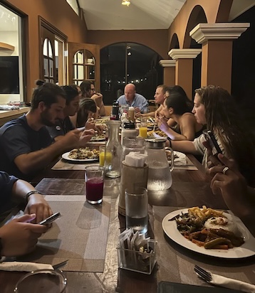 Students and researchers sit at a long table with dinner plates full of food in front of them. Some people are eating, others are talking to each other.