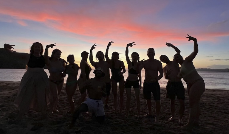 Studetns and researchs stand shoulder to shoulder waving and throwing up peace signs at the camera. They are all covered in shadows while a beautiful sunset with pink clouds sits behind them.