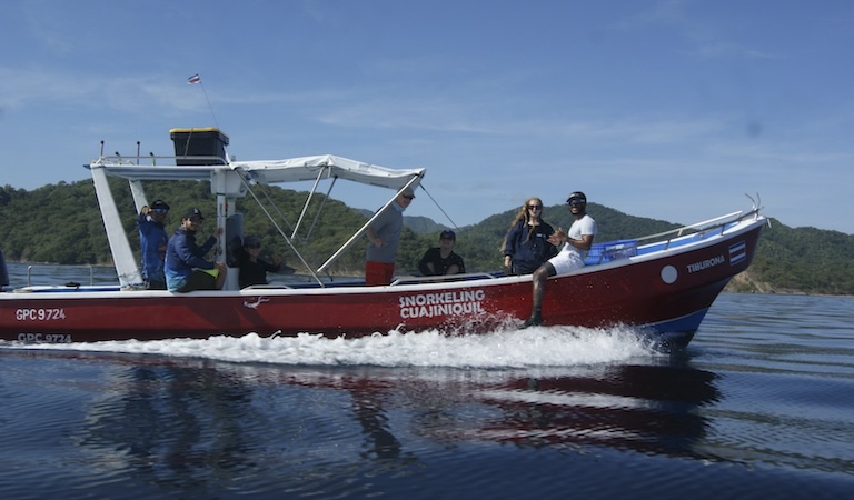 Students and researchers smile and wave at the camera. They're on a red boat with white trim that is sailing on water.