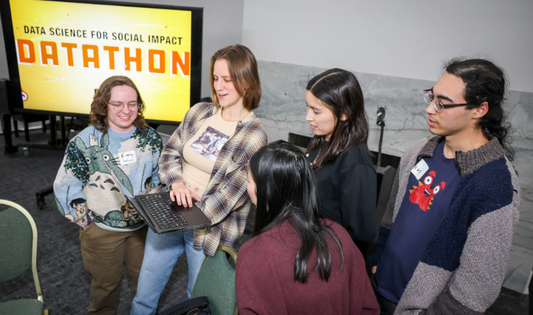 Students look surprised and intrigued at a laptop. A monitor in the background reads, "Data Science for Social Impact Datathon."