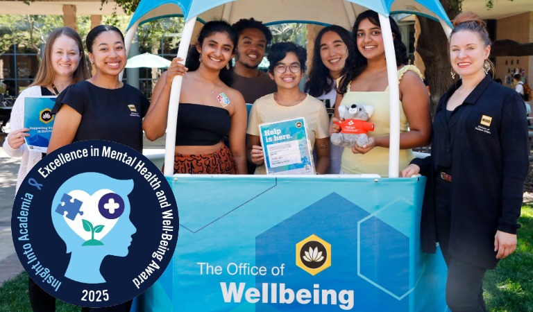 Students and staff from the Division of Student Affiar's Office of Well Being pose for a picture. They are all smiling and a few of them are holding Office of Well Being branded flyers that read, "help is here." The seal for the 2025 Excellence in Mental Health and Well-Being Award is fixed to the bottom left corner of the picture and includes the soft blue side profile of someone's head imposed with a white heart. Inside the white heart are icons of a puzzle piece, a medical cross, and a sprouting plant.