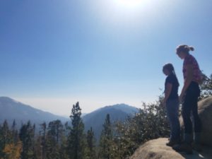 Two students stand on a boulder as they look off into the tree covered canyon. In the background, the mountains are outlined and show off various shades of blue and green. Above, the sky is completely clear allowing for the solar rays to penetrate all parts of the wilderness.