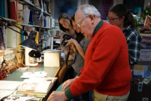 An older man wearing a red sweater and three young women look down at a book in a professor’s office