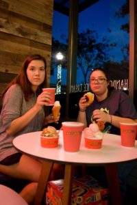 Two girls sit at a table and stare into the camera, surrounded by three cups of coffee and four types of ice cream.