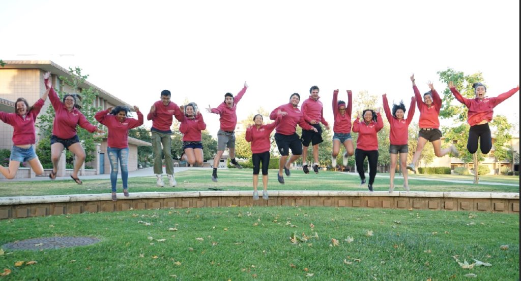 Fourteen college students on a lawn wearing identical red sweatshirts jump in the air