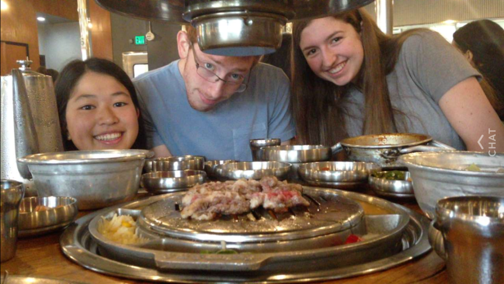 Three students with many metal bowls and meat on a Korean barbecue grill.