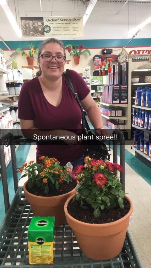 A smiling girl pushes a cart carrying two potted flowers and a container of fertilizer