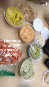 One bag of carrots and multiple containers of different types of hummus and goat cheese are spread out on a table. The caption reads “Hummus and goat cheese galore!”