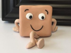 An anthropomorphic square prism smiles and crosses its legs, appearing to be sitting down