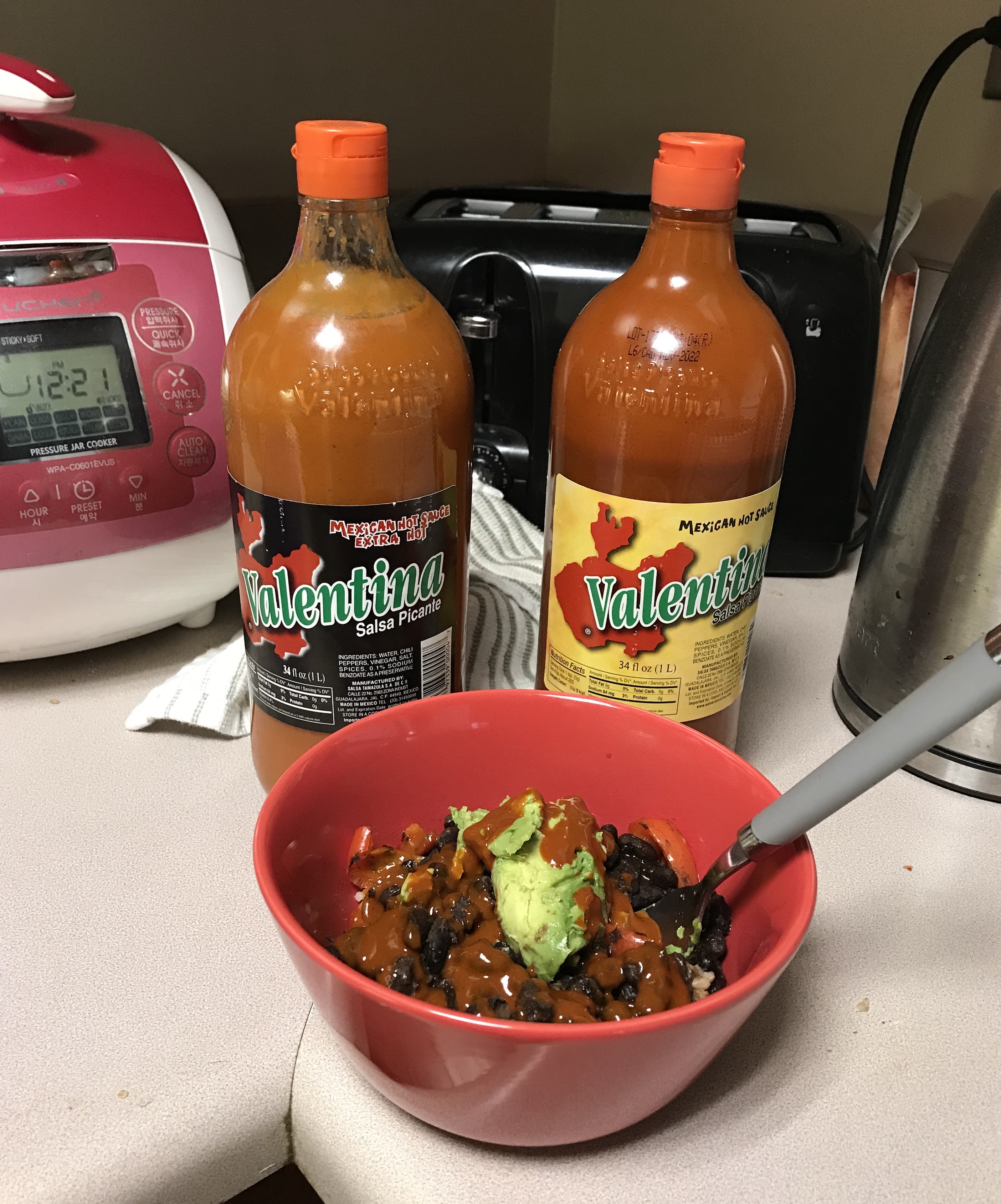 A bowl of black beans, rice, avocado, and Valentina hot sauce sitting in front of two extra large bottles of Valentina hot sauce
