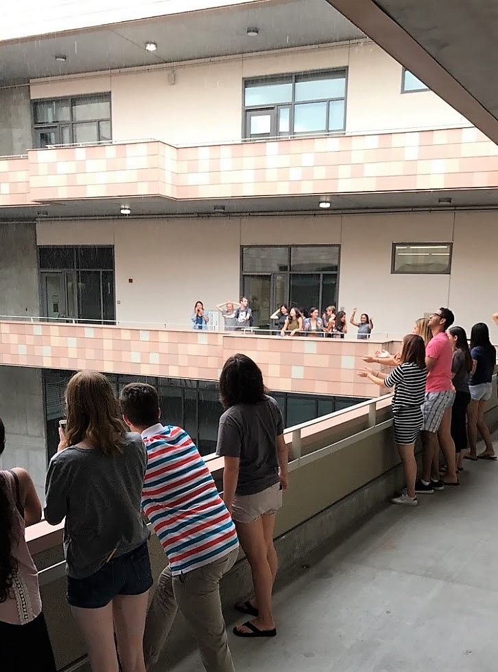 A group of students standing on the balcony of an academic building staring at the sky where rain is falling.