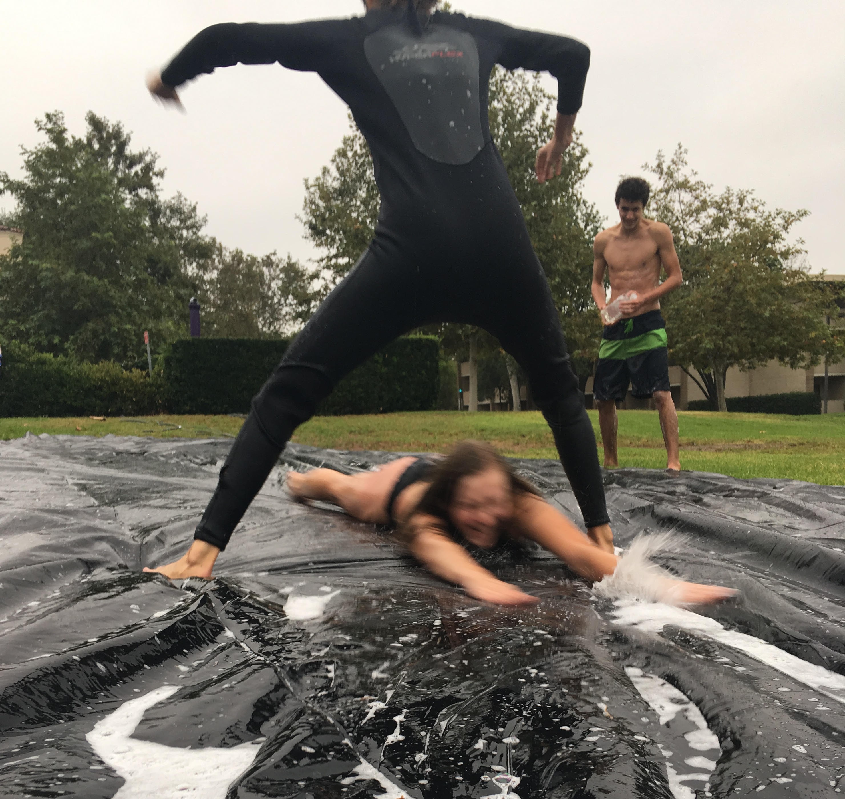 A girl sliding on her stomach on a tarp through the legs of another girl standing in a westsuit.