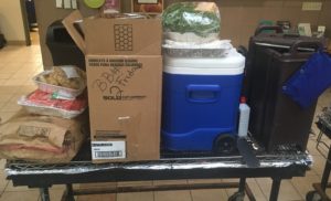 Picture shows a grill with lots of items stacked on it, including a large tray of fruit, a cooler for meat patties, a box of chips and plates, and a cooler/dispenser for lemonade.
