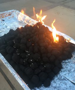 Picture shows coals on a tin-foil surface, on fire. The coals are mostly black, with some spots of gray.