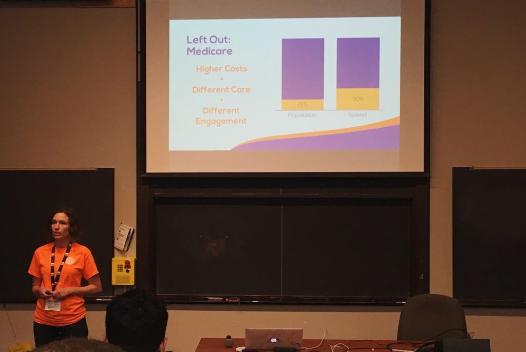 A woman stands in front of a slide that says: "Left out: Medicare. Higher Costs, Different Care, Different Engagement" There is a graph showing that Medicare is 15% of the population but 30% of the healthcare spending.