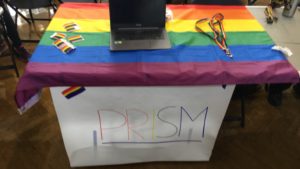 Pictured is a poster with the word PRISM written on it, hanging off a table, with a rainbow pride flag covering the table.