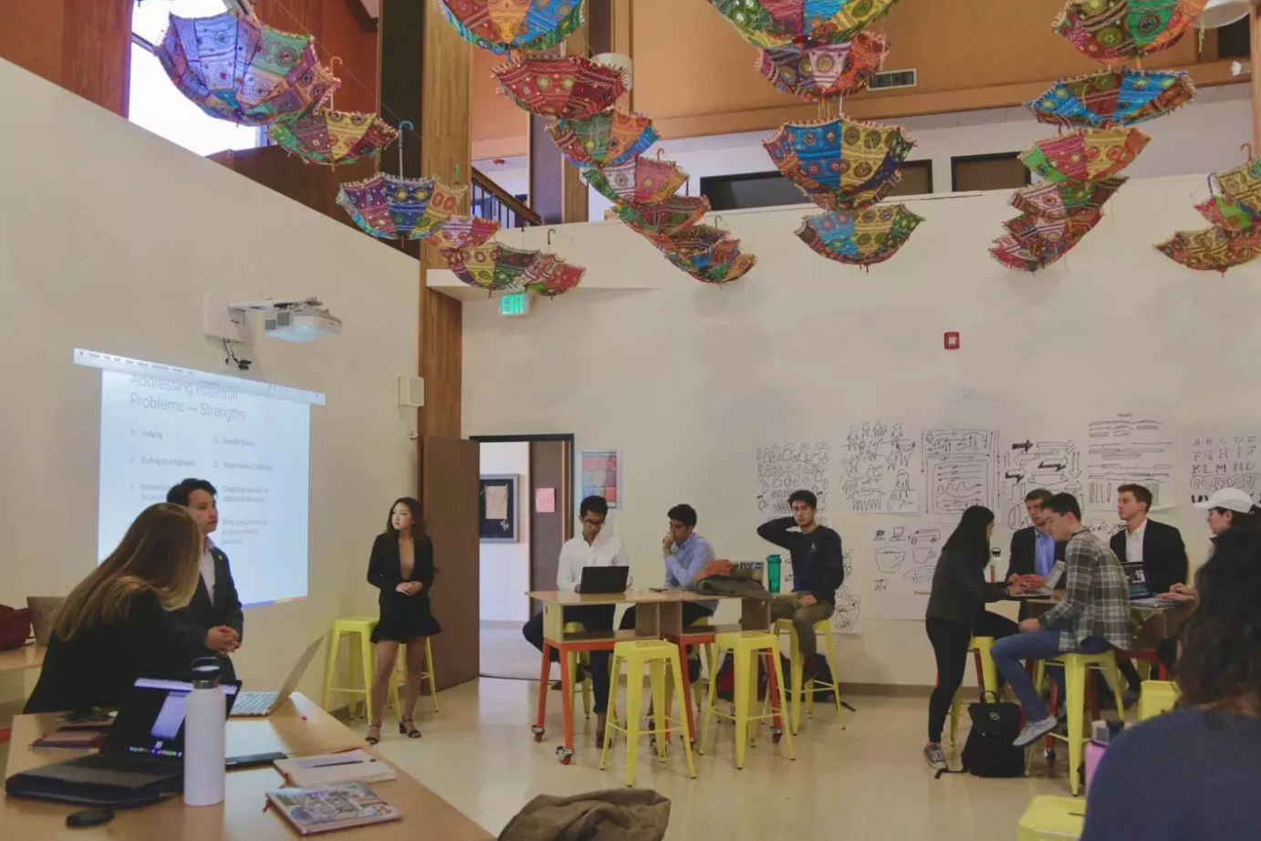 Students are listening to somebody give a presentation at the Hive. There are colorful umbrellas hanging from the ceiling, and people are sitting in groups at tables