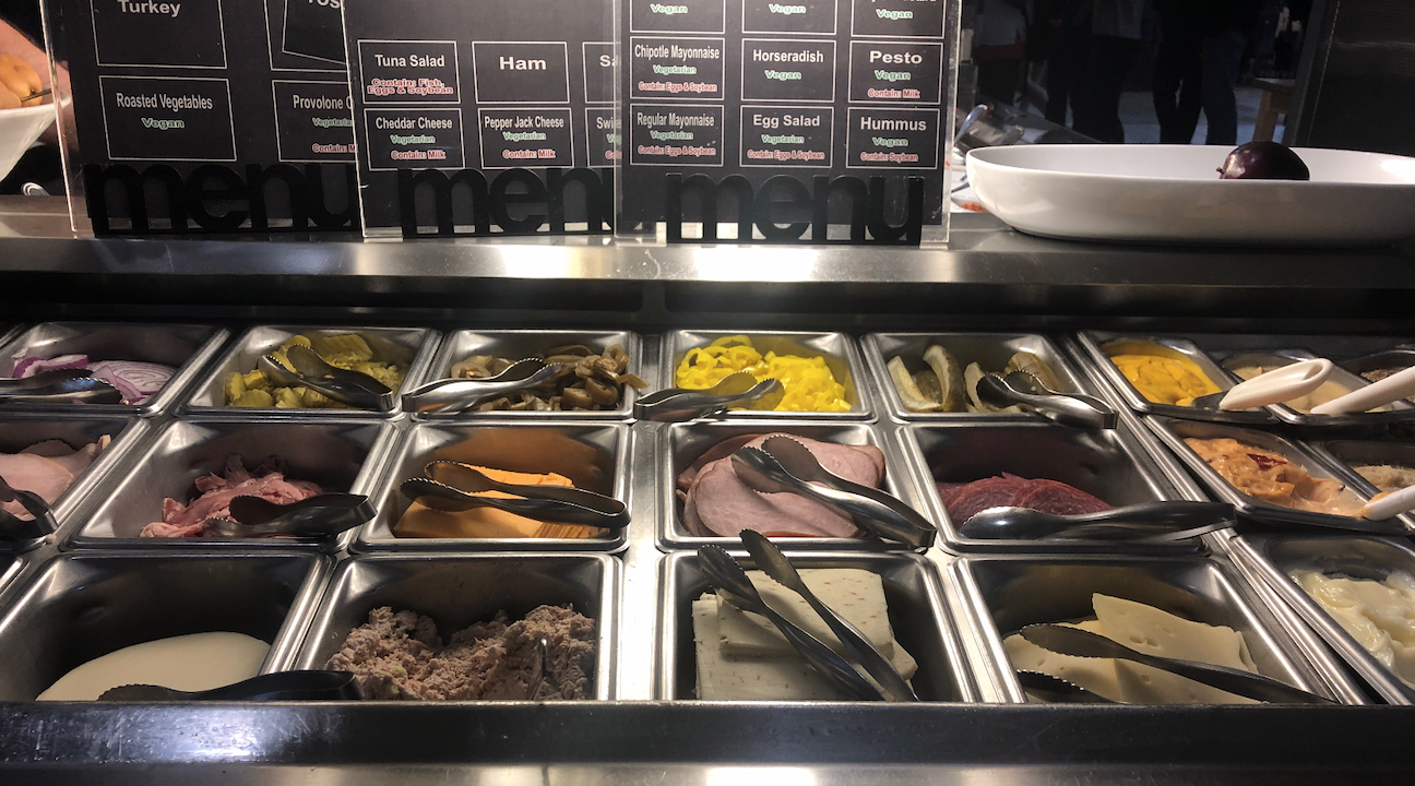 The sandwich bar has tuna, cheese slices, onion slices, roasted veggies, banana peppers, more sliced meat, and spreads like hummus and mustard
