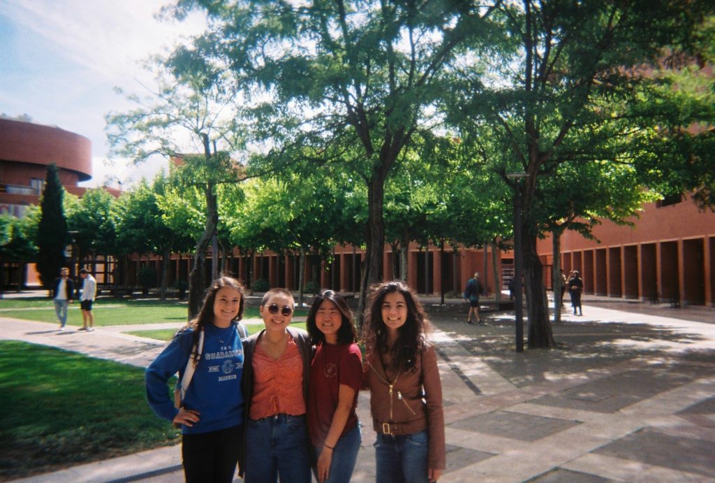 four girls in front of a lawn, trees, and a brown building