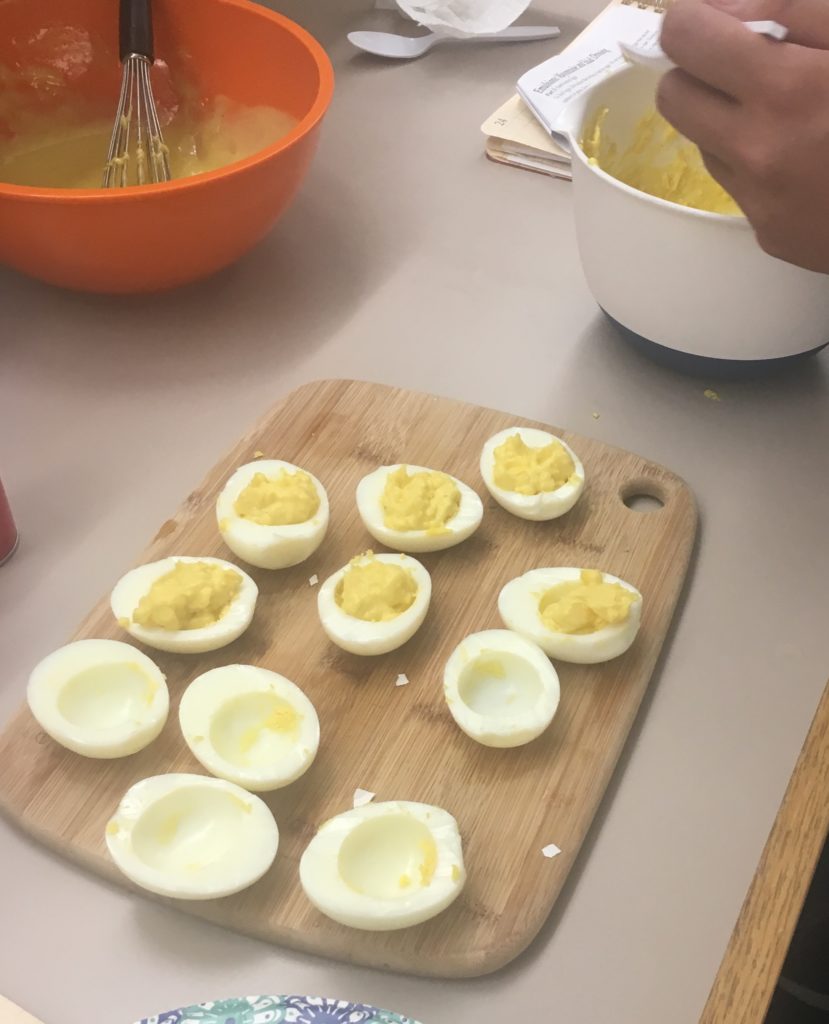 An image showing how my partner and made our deviled eggs. Several filled and unfilled boiled egg whites sit on a cutting board while my partner fills them.