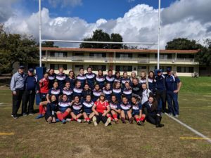 Photo of the 2018-2019 Women's Rugby team. There are 32 players and 5 coaches. everyone is wearing a blue uniform except for two player and a coach. The coach is wearing a grey sweater and the two players are wearing red jerseys.