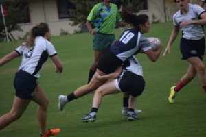 There are four players and a referee. The referee is wearing a light green shirt, three of the players are wearing white jerseys while one is wearing a navy blue jersey. Two of the players in white are at opposite ends of the photo. The one closest to us has her back turned and has a number 3 on her jersey. the third player in white as well as the player in blue are in the center. The blue player is carrying the ball and 19 is sen on her back, while the white player tackles her mid-air. The referee is watching in the background.