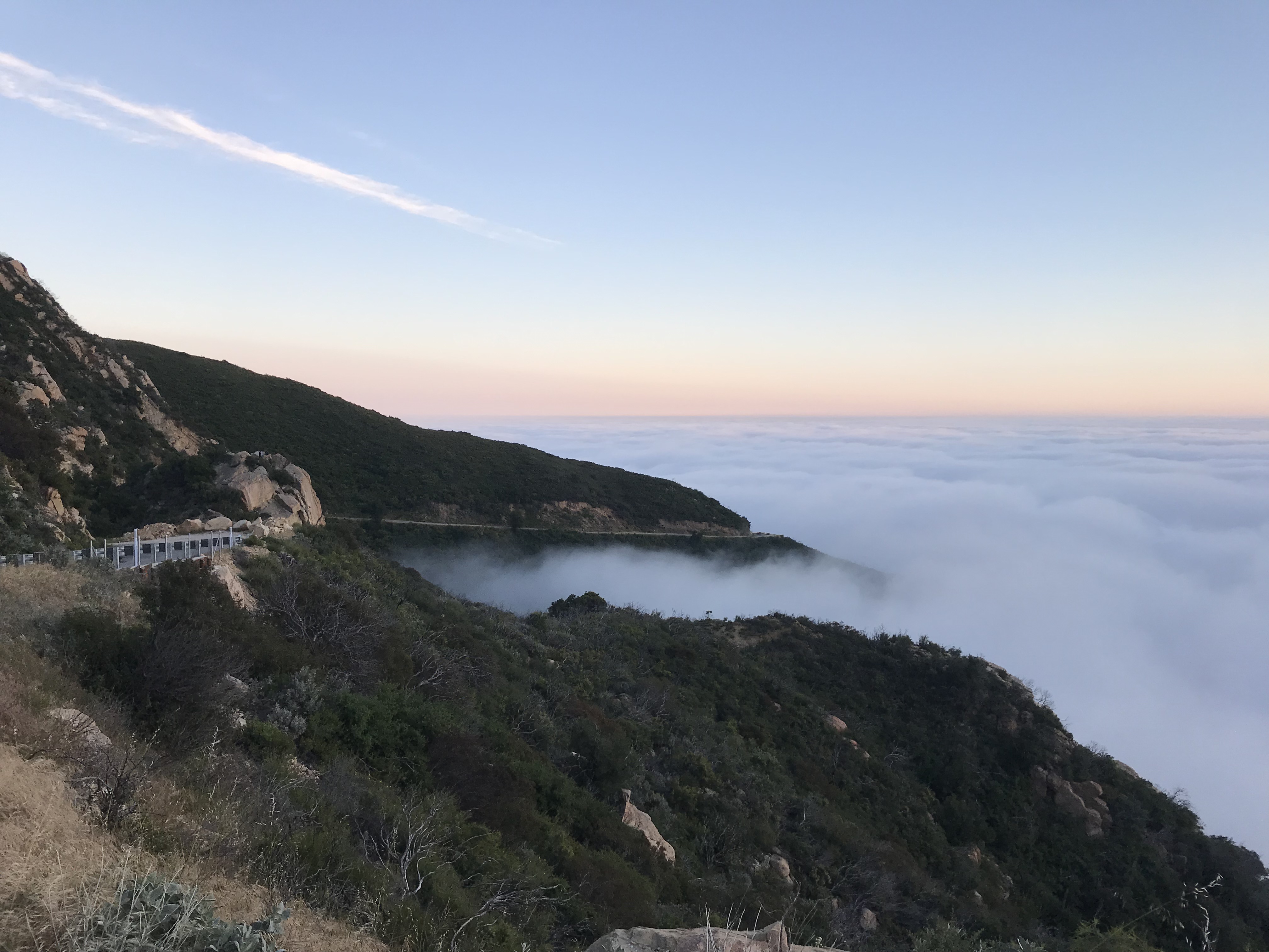 The view from the side of the road near the top of a mountain. The view is above the clouds, which creep into the mountainside. The sky is slightly orange from the sunset.