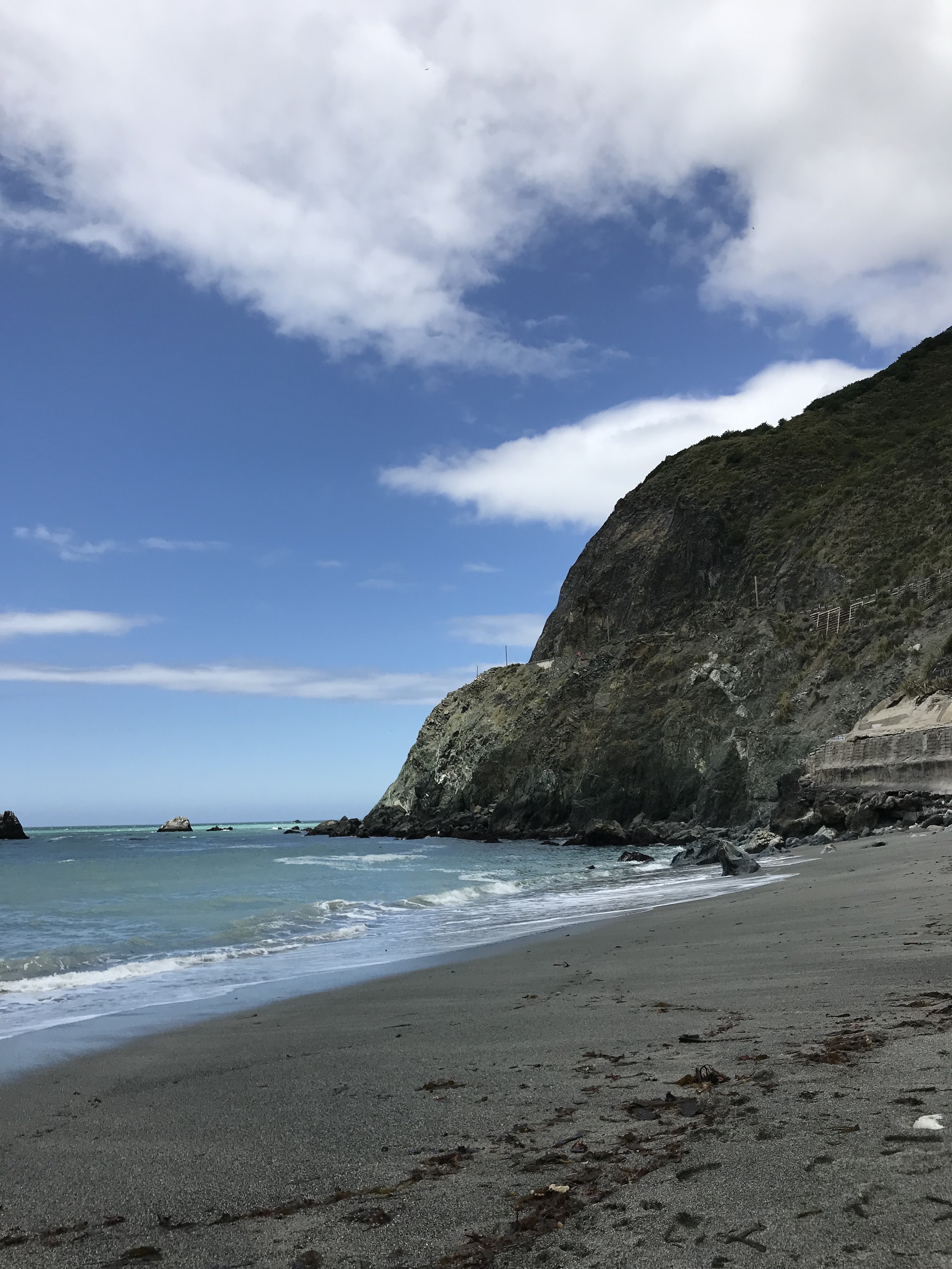 The beach at Big Sur. Tall cliffs are on the right side, and the sky is super blue with a couple of white clouds. The sand is a dark color from the pebbles, and the water is pretty calm.