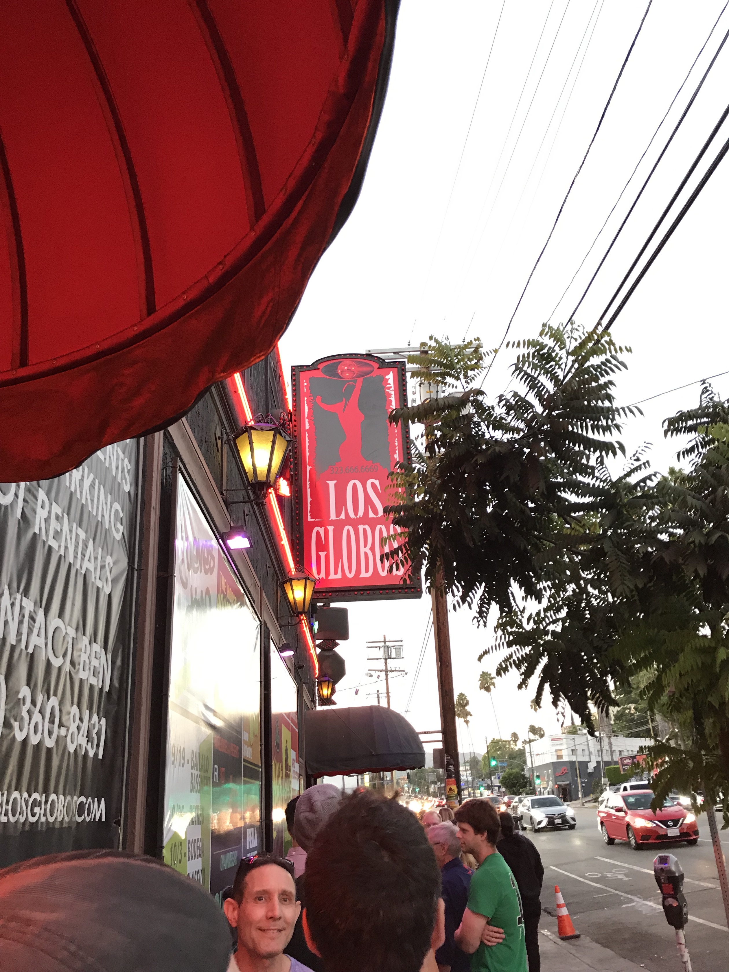 A large red sign says "Los Globos" and is hanging from the side of a building. There is a line of people waiting to get into the door, which is also on the side of the building. One person in the line has their face to the camera and is smiling.