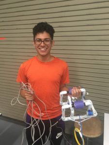 Mason Acevedo, Class of 2022, Standing, holding an underwater robot. In one hand, he has the robot frame, made mostly of PVC pipes. In the other hand are wires connected to the robot.
