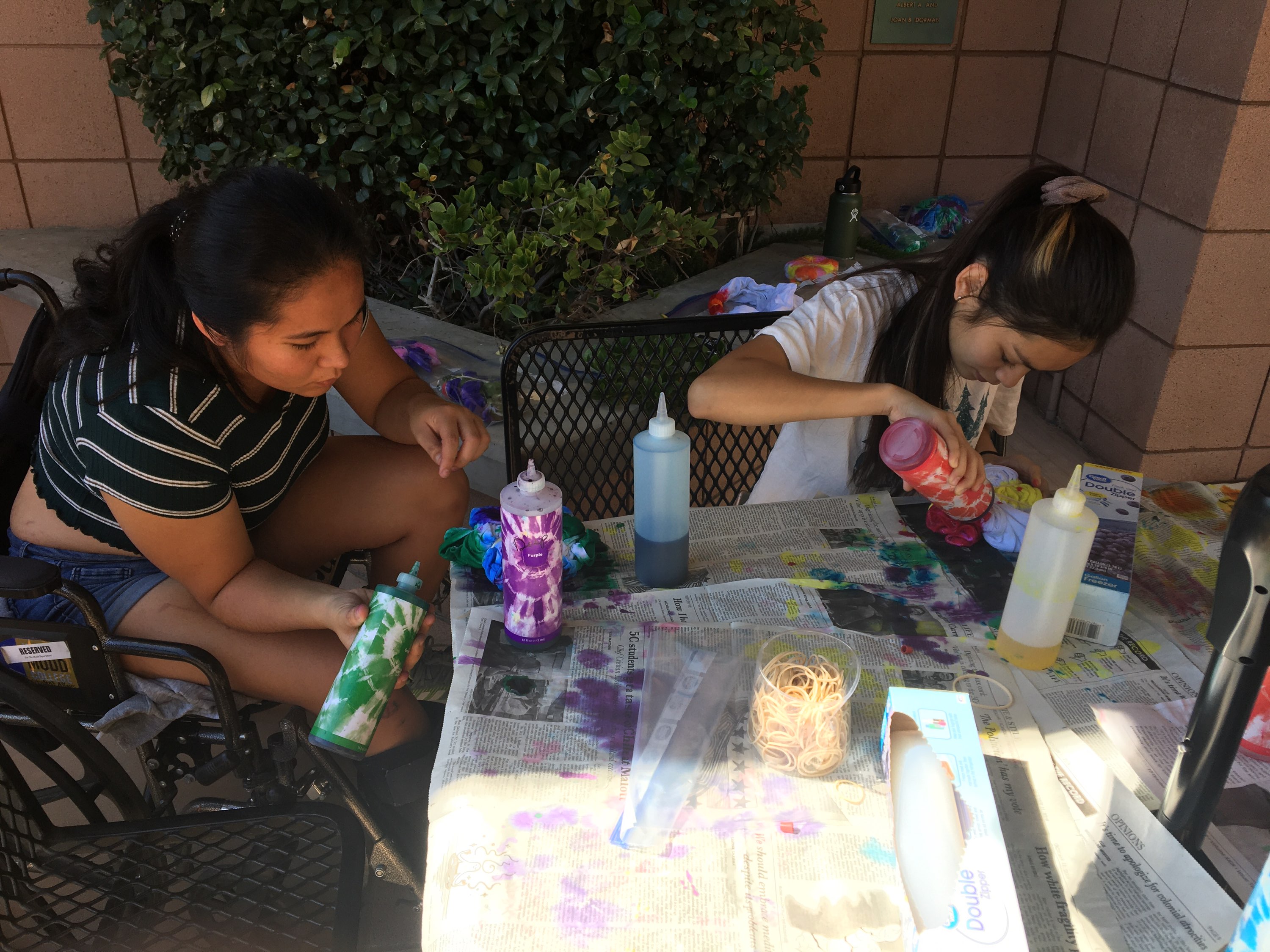 Two Mudd students sitting at a table use bottles of dye on white shirts in front of them.
