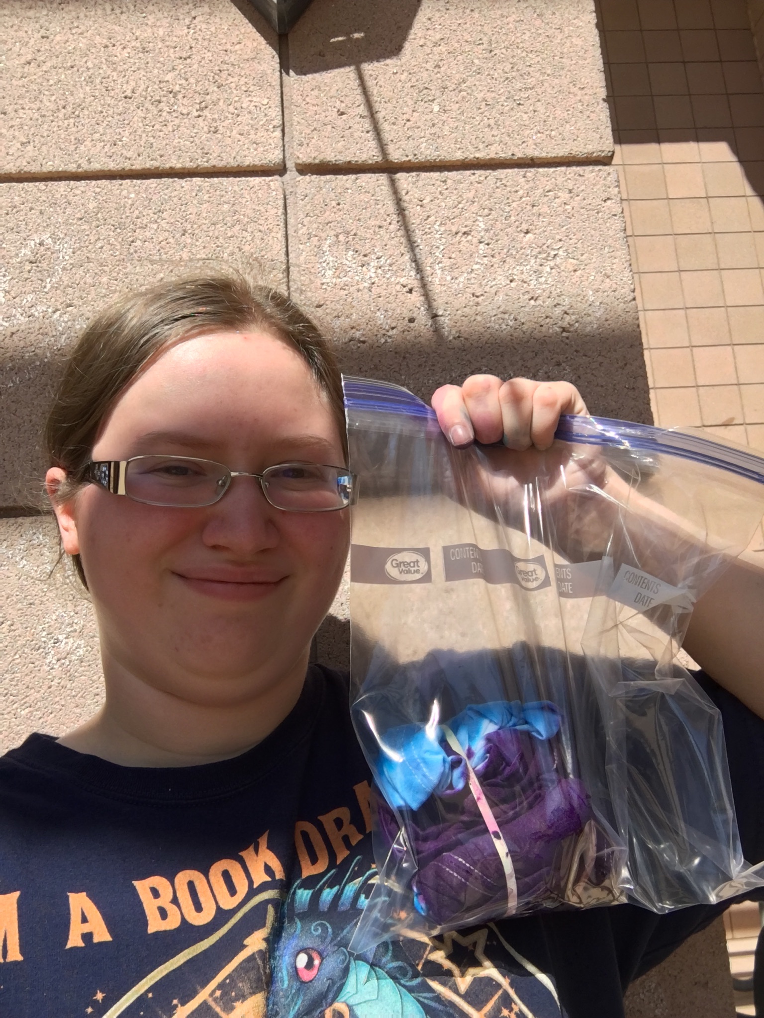 Anya holds up a ziploc bag with a balled-up blue and purple shirt in it.