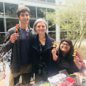 A photo of a group of three wellness peers (with me on the right), at the wellness pop-up in front of the Hoch. Everyone is holding a fruit skewer and either sitting or standing in front of a table full of fruit.)