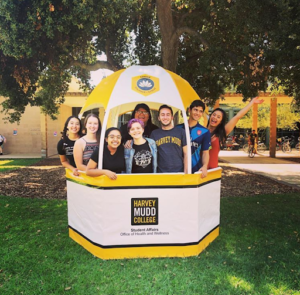 A photo of a group of 8 students inside a white and yellow gazebo with the Office of Health and Wellness along with the Harvey Mudd logo.