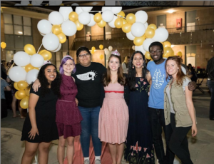 A photo of a group of students dressed nicely with myself near the center left and Dean Michelle on the right, all standing in front of a yellow and white balloon arch.