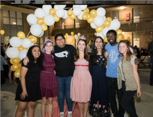 A photo of a group of students dressed nicely with myself near the center left and Dean Michelle on the right, all standing in front of a yellow and white balloon arch. 