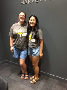 Kailee and Hannah, wearing tour guide shirts, blue shorts, and Birkenstocks, pose with smiles in front of a wall in the Admissions Office.