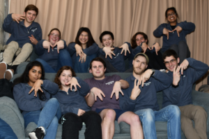 A group of college students sit on two tiers of couches. They are wearing blue sweatshirts (except one person in the middle wearing a purple shirt) and forming Mâs with their fingers in different ways.