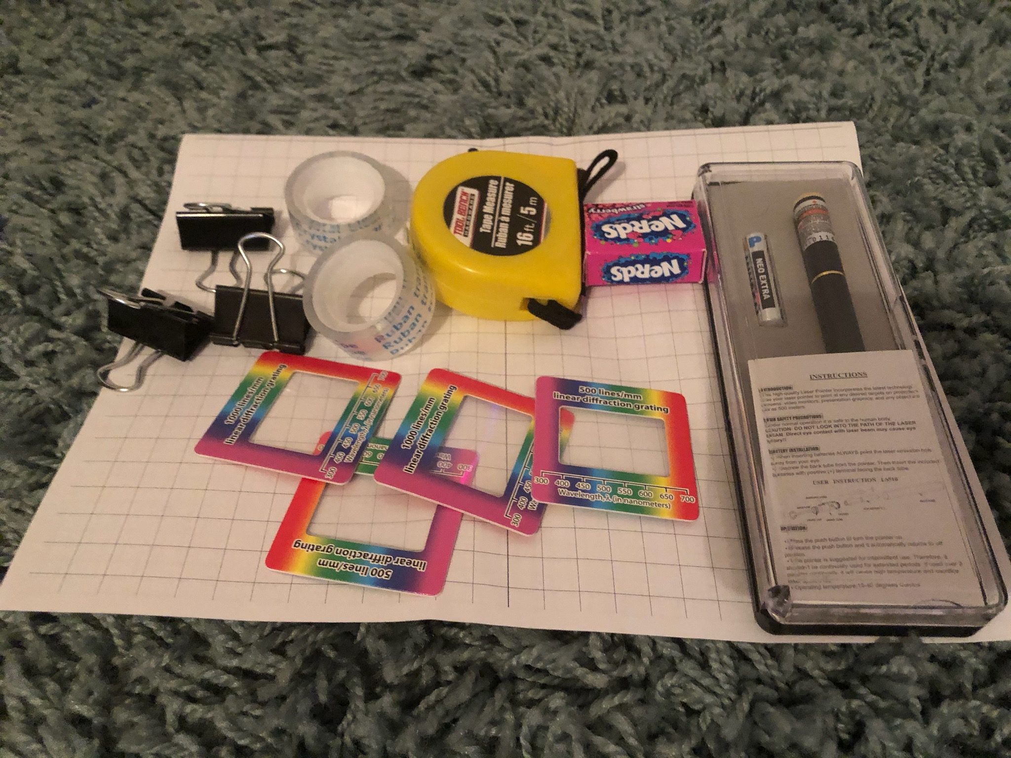 Three binder clips, a tape measure, four filters, two rolls of tape, a laser pointer and a container of Nerds candy on graph paper.