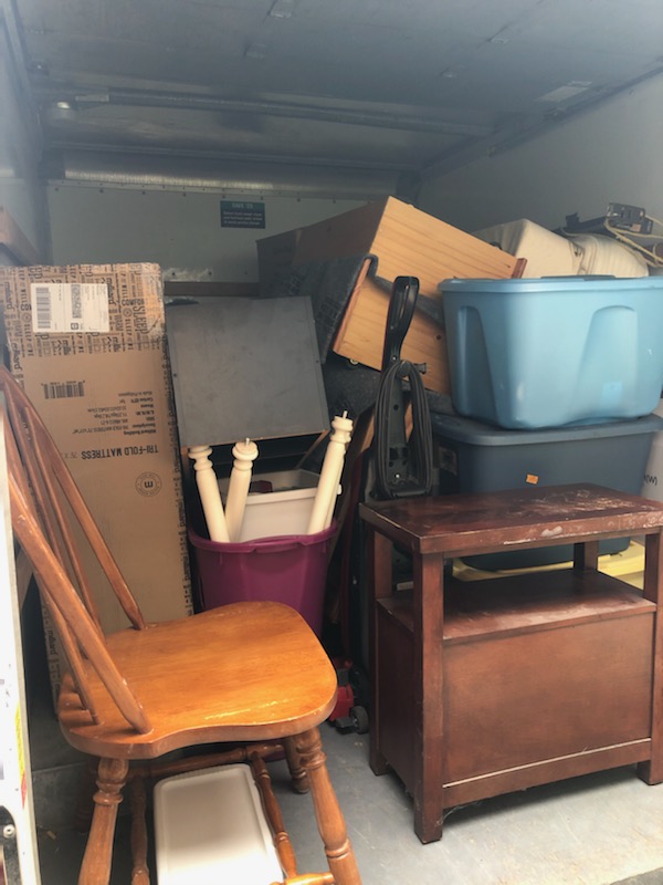 The back of a moving truck, with a chair, a bedside table, and several boxes visible.