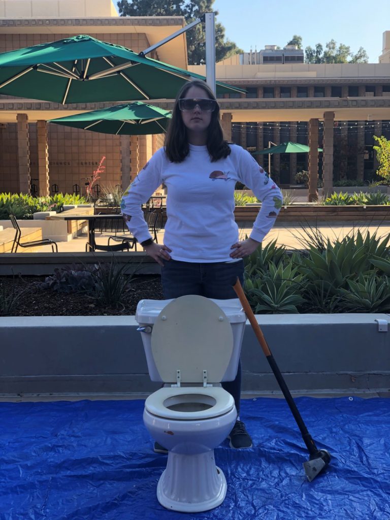 Skylar standing by a toilet with a large hammer