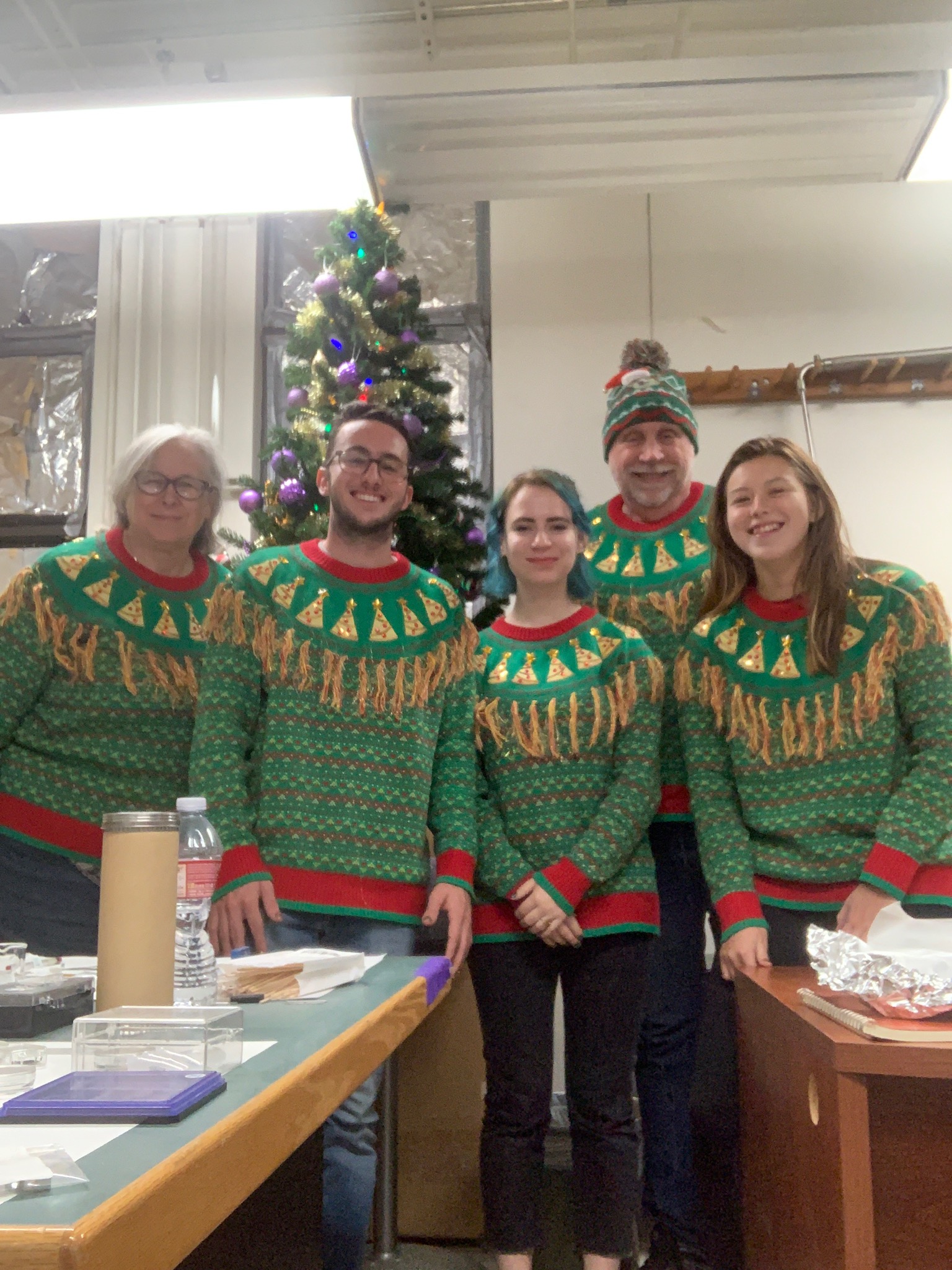 Three students and two professors wearing matching Christmas sweaters in front of a Christmas tree