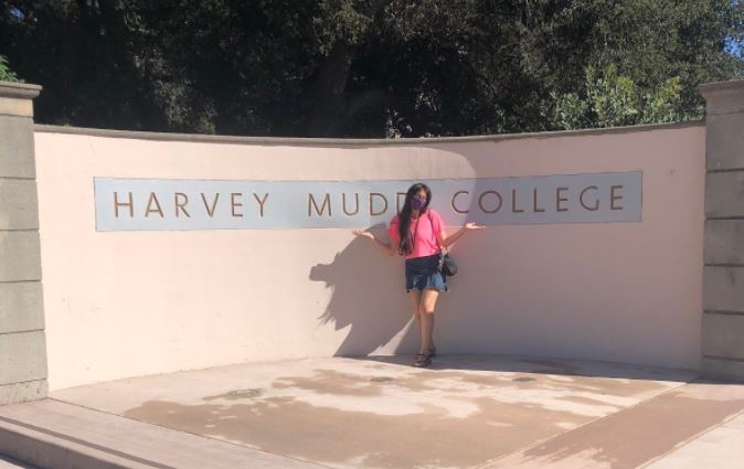 Ana in front of a white and blue sign that reads "Harvey Mudd College"