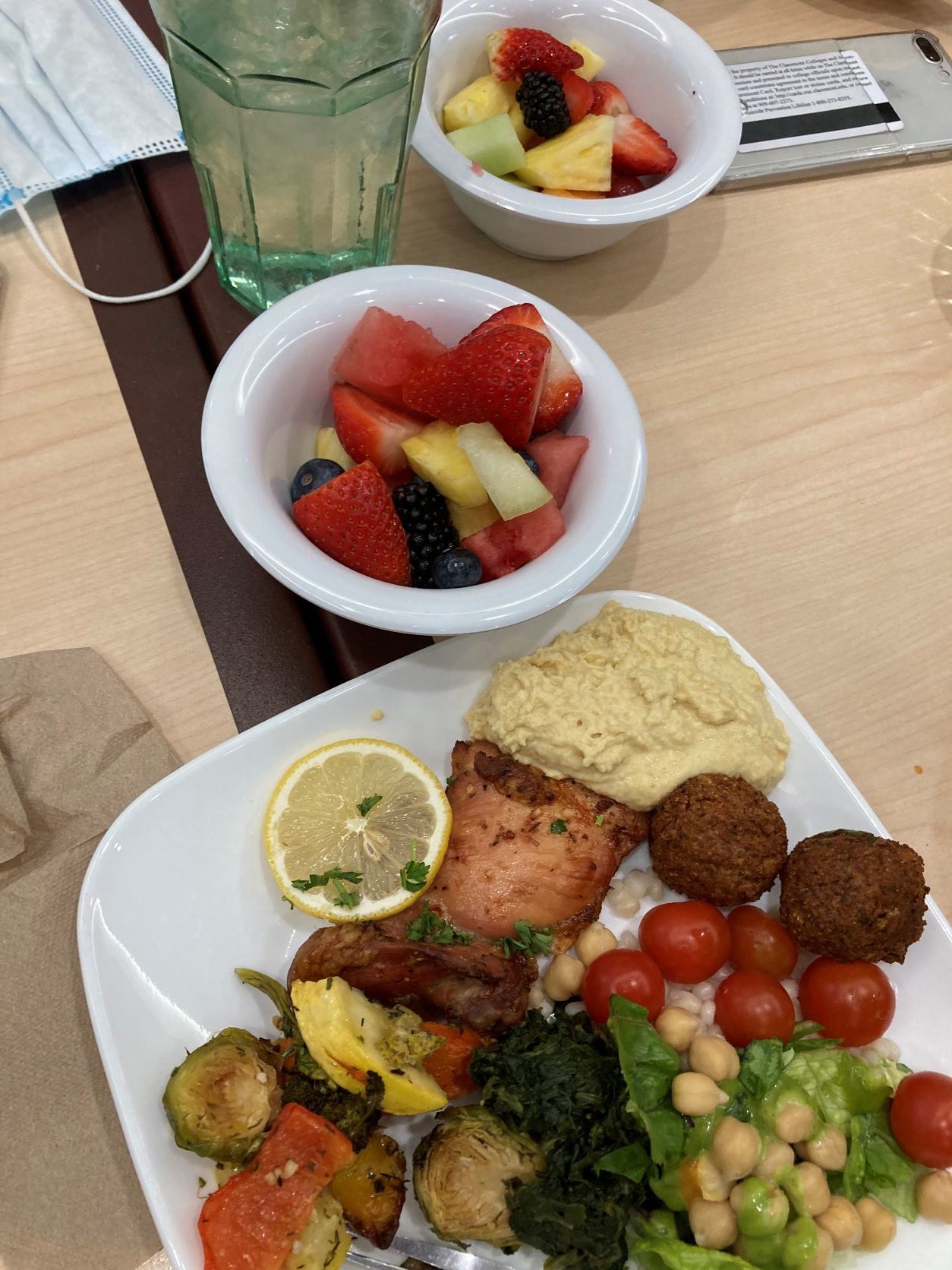 A small bowl of fruit next to a plate with a selection of salmon, bread, vegetables, and falafel
